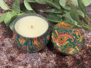 Decorative candle with paisley design lid on a textured surface with green leaves.