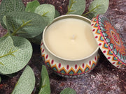 Decorative candle in a colorful tin with a patterned lid, surrounded by green leaves.