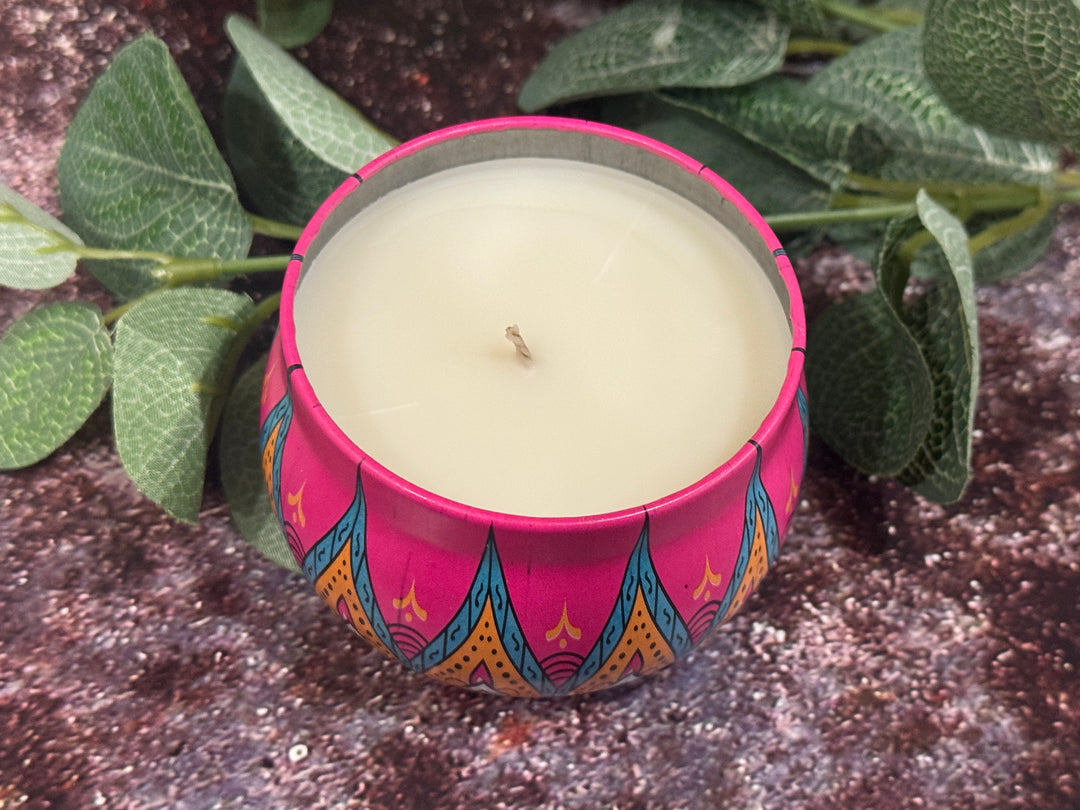 Candle in a colorful pink tin jar on a dark surface.